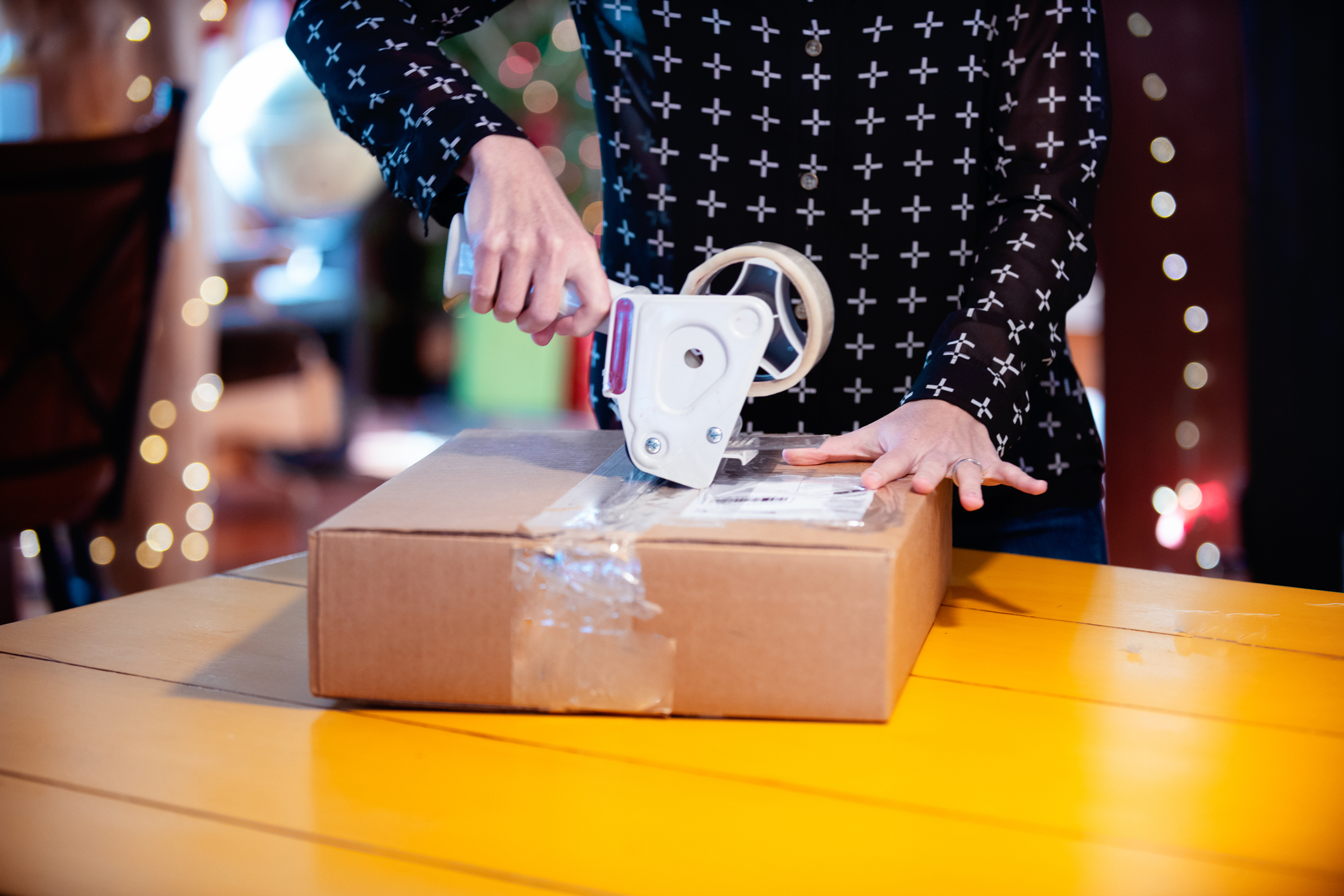 A box or package on a table. There is Christmas decoration in the background. Adult is taping the container with a tape tool. The box is closed with adhesive tape. Mother returns the dress by parcel. Photo was taken in Quebec Canada.