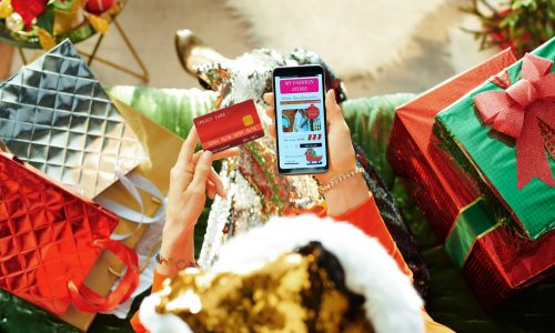 Holiday season. female in sparkle gold sequin santa hat with credit card browsing high fashion retail online store and on a smartphone while sitting on divan in the modern living room at Christmas.