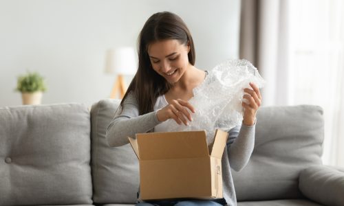 Happy young woman open cardboard box satisfied with purchase online shop order sit on sofa at home, smiling lady customer receive unpack parcel look inside, postal shipping courier service concept