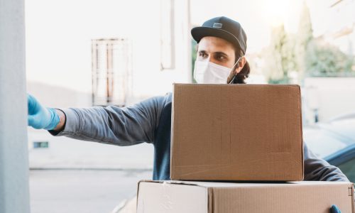 Delivery man is ringing the bell to deliver two cardboard boxes. He's wearing a face mask and protective gloves.