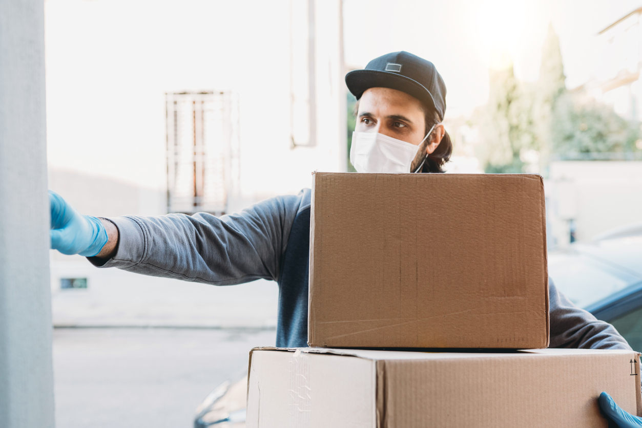 Delivery man is ringing the bell to deliver two cardboard boxes. He's wearing a face mask and protective gloves.