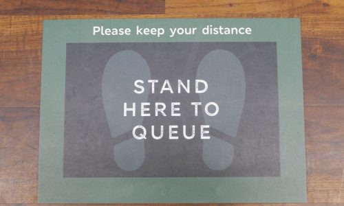 A food store queue instruction floor sticker, stuck to the wooden floor of the shop, during the Coronavirus pandemic. Customers are asked to stand two metres apart, using the footprint floor sticker as a distance marker within the store, while waiting near the checkout tills.