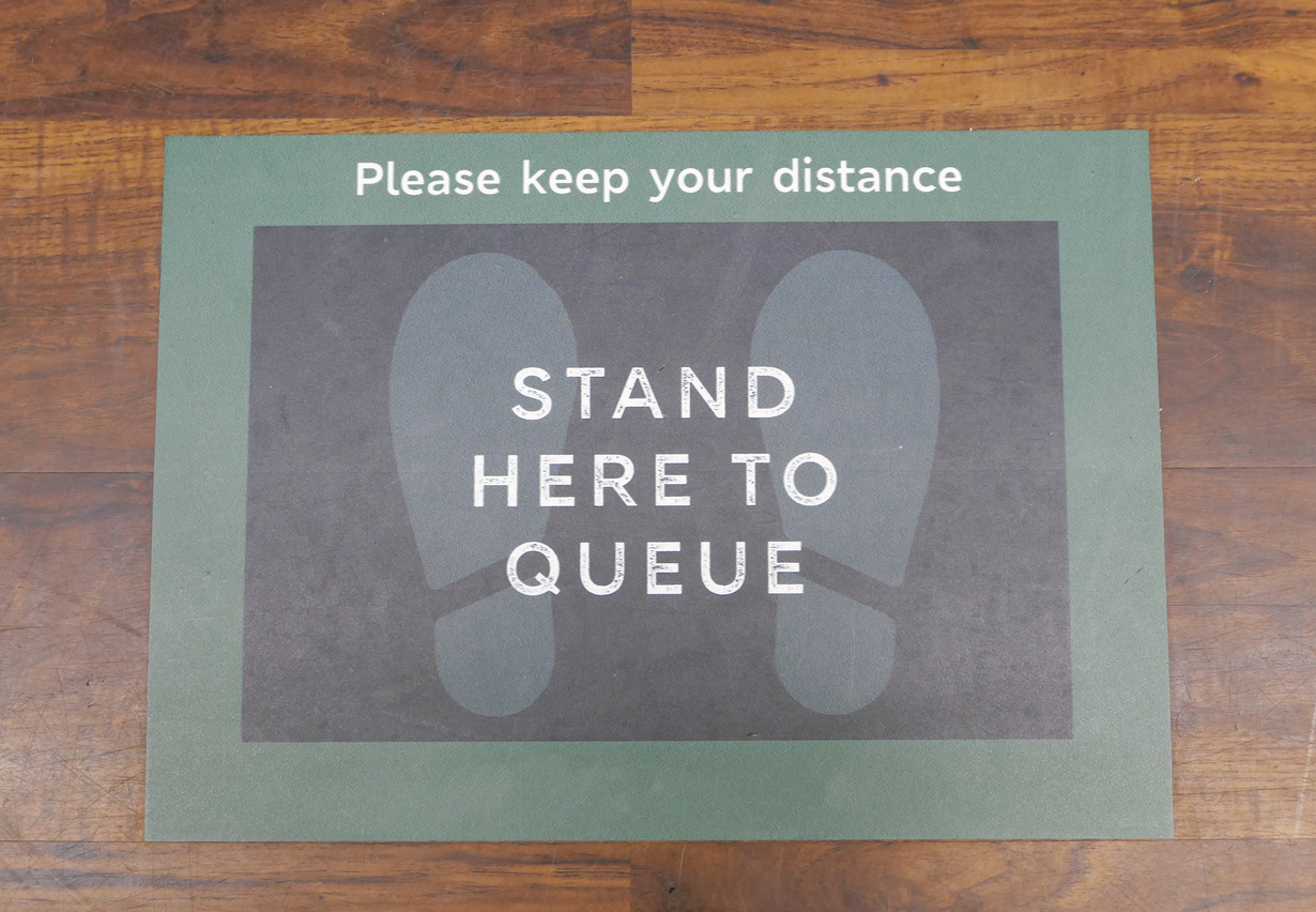 A food store queue instruction floor sticker, stuck to the wooden floor of the shop, during the Coronavirus pandemic. Customers are asked to stand two metres apart, using the footprint floor sticker as a distance marker within the store, while waiting near the checkout tills.