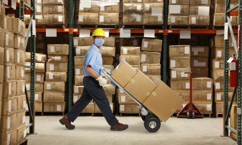 A warehouse worker wearing a protective mask and a hard hat pushes a hand truck and a stack of boxes in a warehouse stacked with inventory.