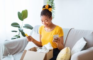 Online Shopping And Payment. Portrait of excited african american female buyer holding credit card and using mobile phone, sitting on couch and looking in cardboard box container with white sneakers