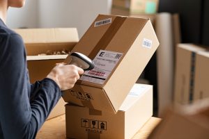 Hands of young woman scanning barcode on delivery parcel. Worker scan barcode of cardboard packages before delivery at storage. Woman working in factory warehouse reading and scanning labels on the boxes with bluetooth barcode scanner.