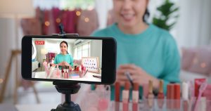 Asian woman records live video at home studio. Beauty products on a table with blurry background.