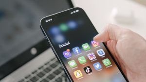 Hand holds an iPhone with social media apps, against the blurred background of a laptop on table.