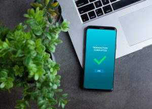 Transaction completed notification in a mobile phone screen, phone alongside with potted plant and laptop on dark stone background