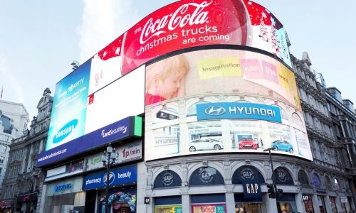 "London, UK - December 2, 2012: People walking and window shopping below the electronic advertising displays of Piccadilly Circus in London. Piccadilly Circus used to be surrounded by illuminated advertising hoardings on buildings since the early 1900s, but only one building now carries them."